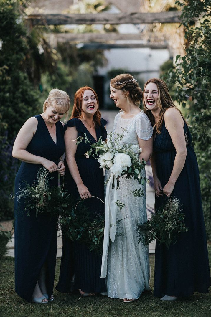 Bride holds white floral bouquet and stands with her bridesmaids in black bridesmaid dresses in differing styles outdoors 