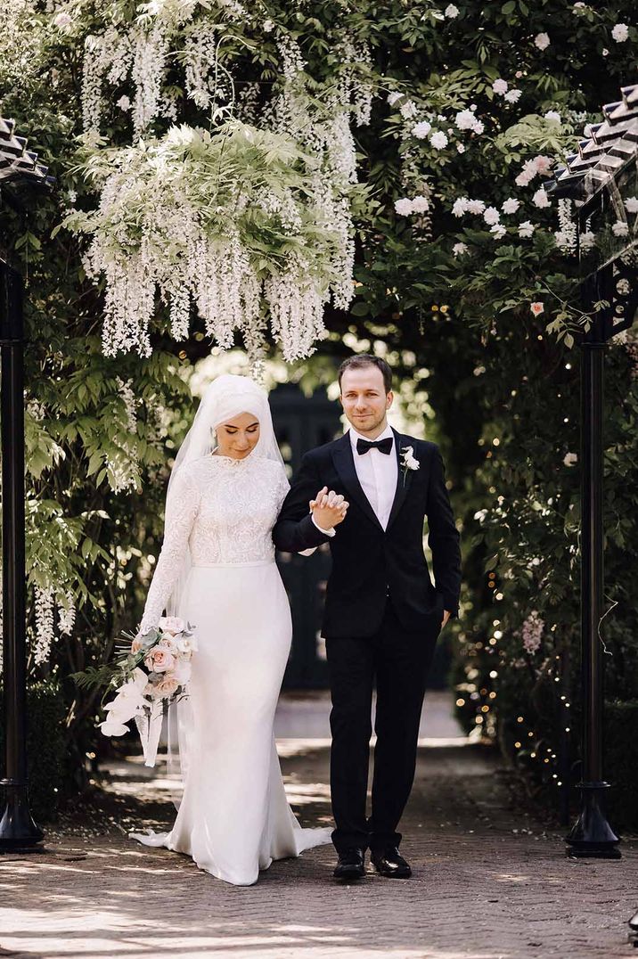 Bride in long sleeve fitted lace wedding dress holding pastel toned floral bouquet and groom in classic black tux walking through wedding venue grounds 