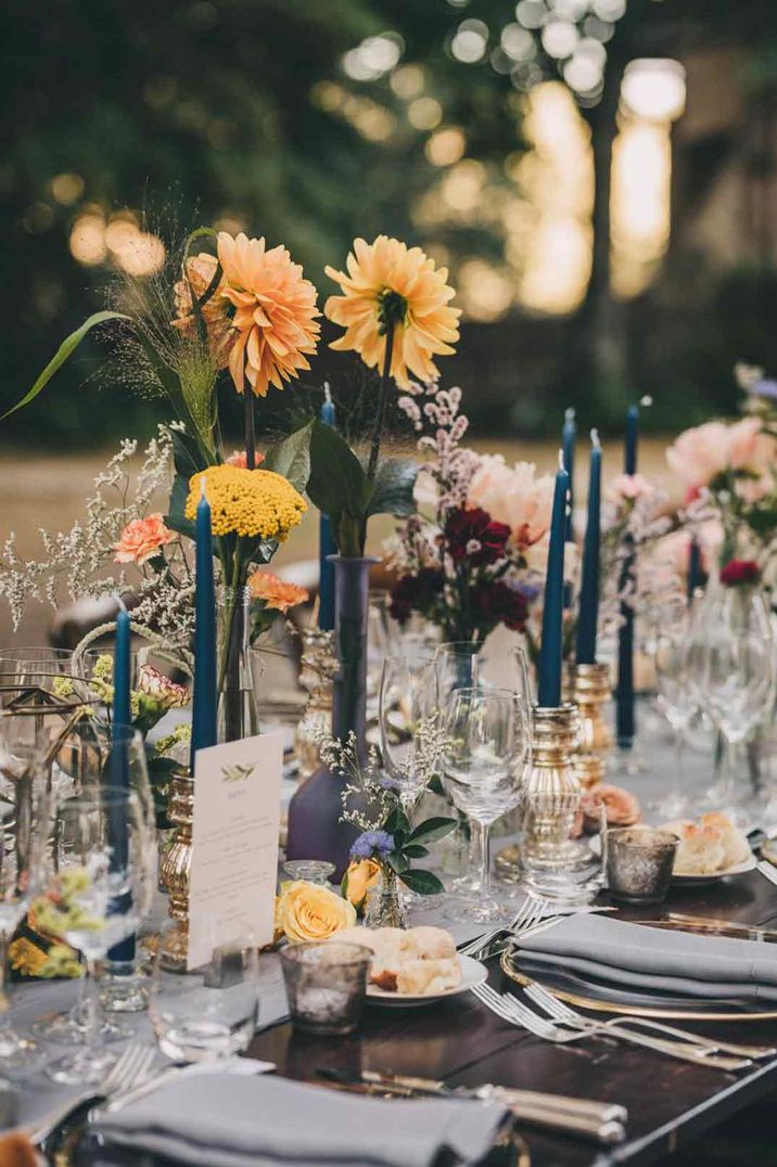 Blue tapered candles and yellow flower stems in vases and bottles on rustic, natural wedding tablescape 