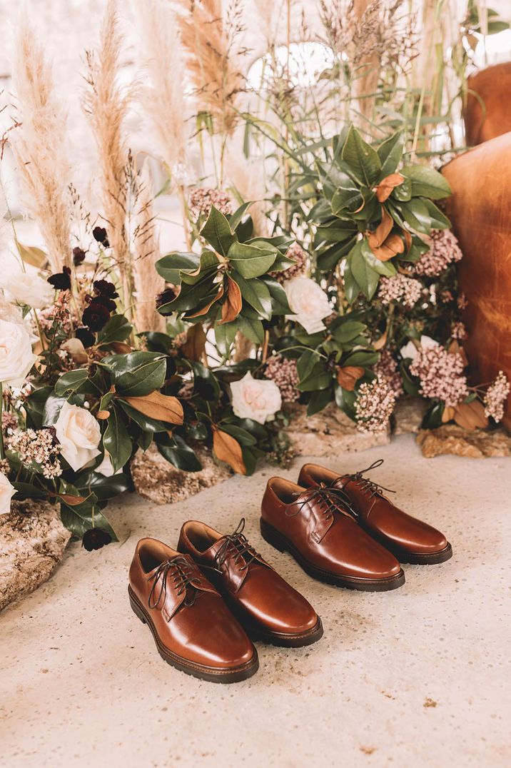 Two pairs of shiny brown leather groom shoes next to pampas grass and white flower altar decoration 