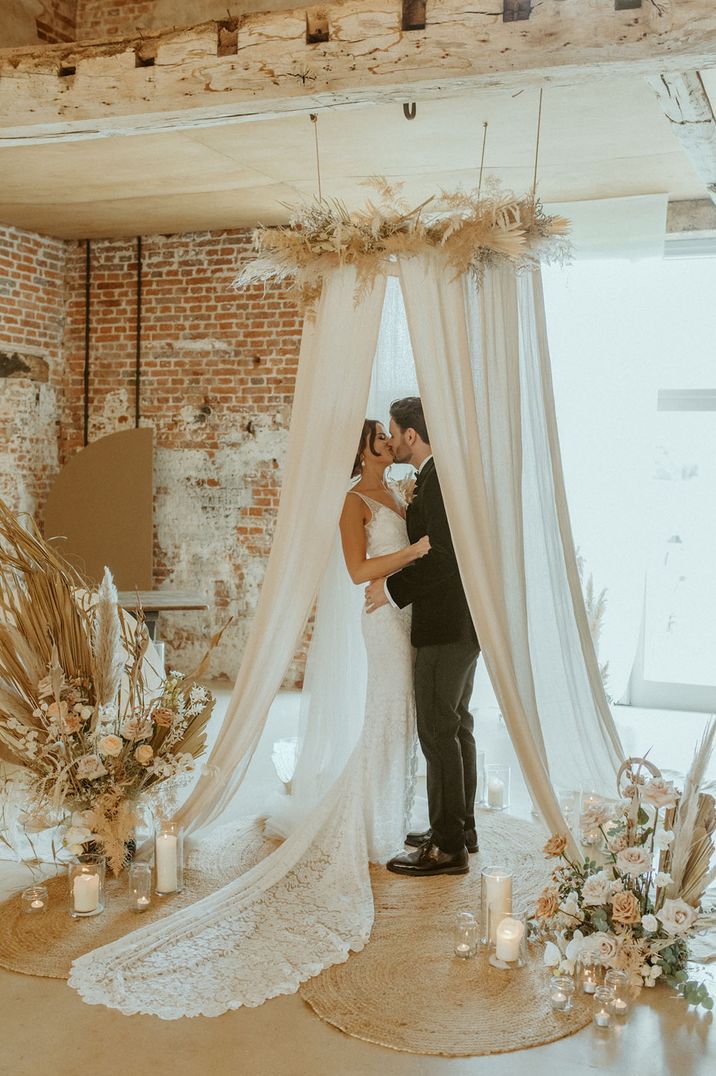 The bride and groom kiss inside white drapery altar decoration with dried flowers 