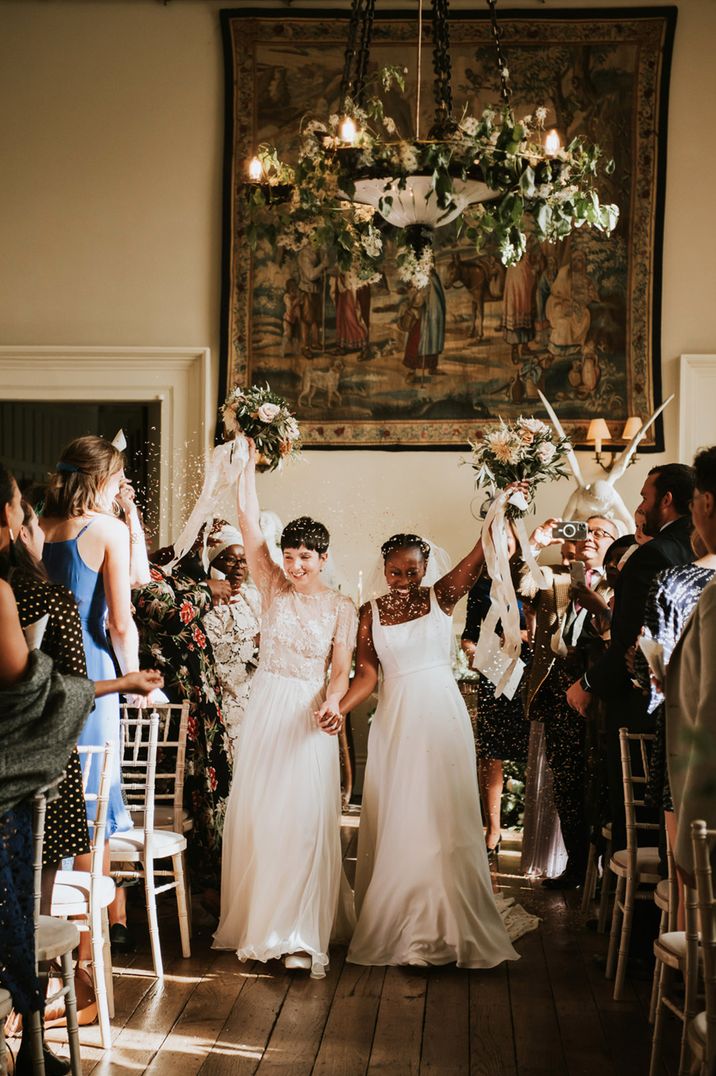 LGBTQI+ wedding ceremony at Elmore court with two brides descending up the aisle as wife and wife waving their bouquets in the air 