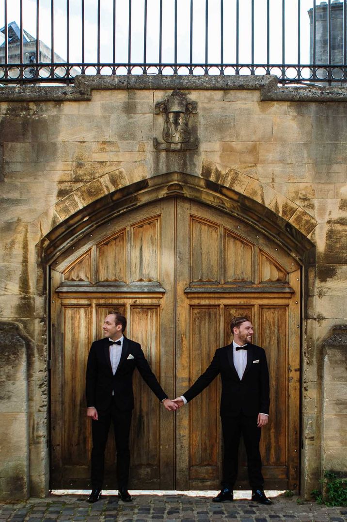 Grooms stand outside of wooden door and hold hands after wedding ceremony 