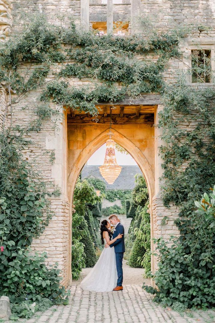 Bride and groom kiss under archway outdoors with a chandelier above at their traditional wedding