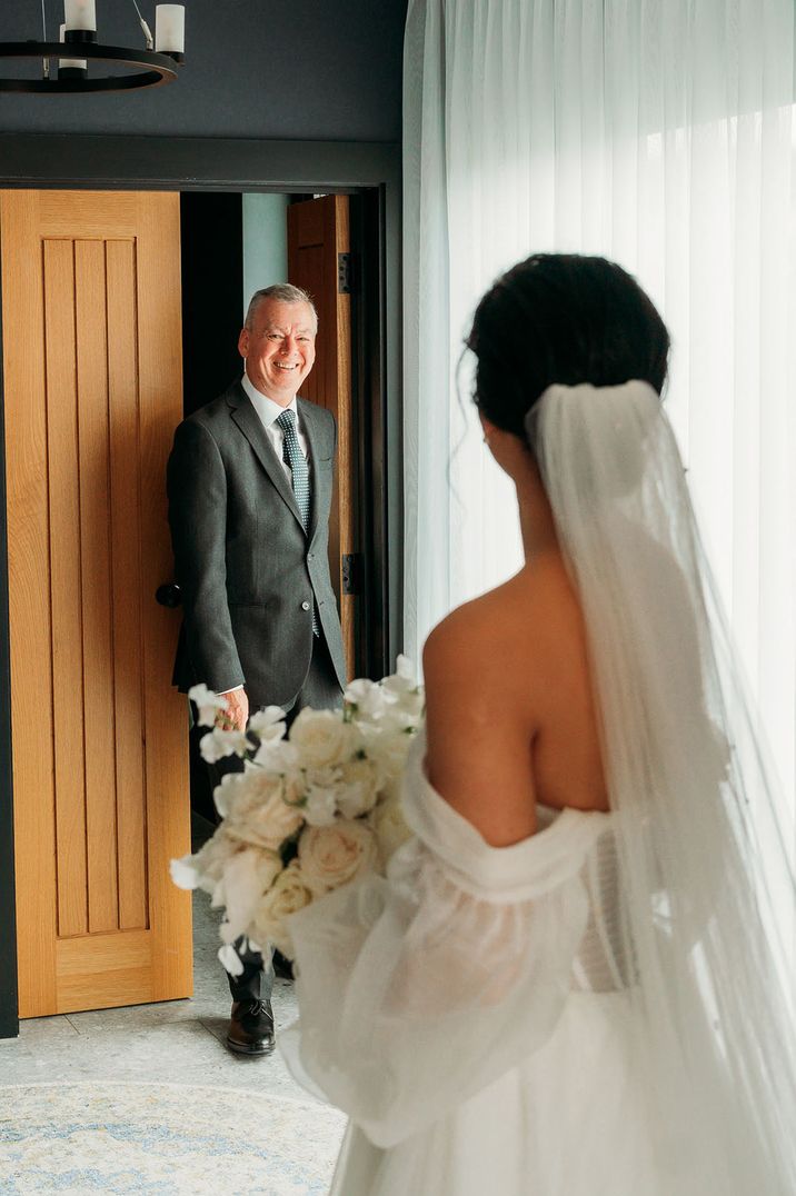 Father of the bride in a grey suit smiles brightly as he spots the bride in her wedding dress 