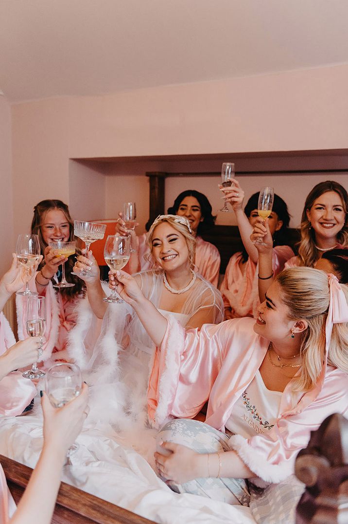 Group of girls in pink pyjamas sitting on a bed together drinking champagne 