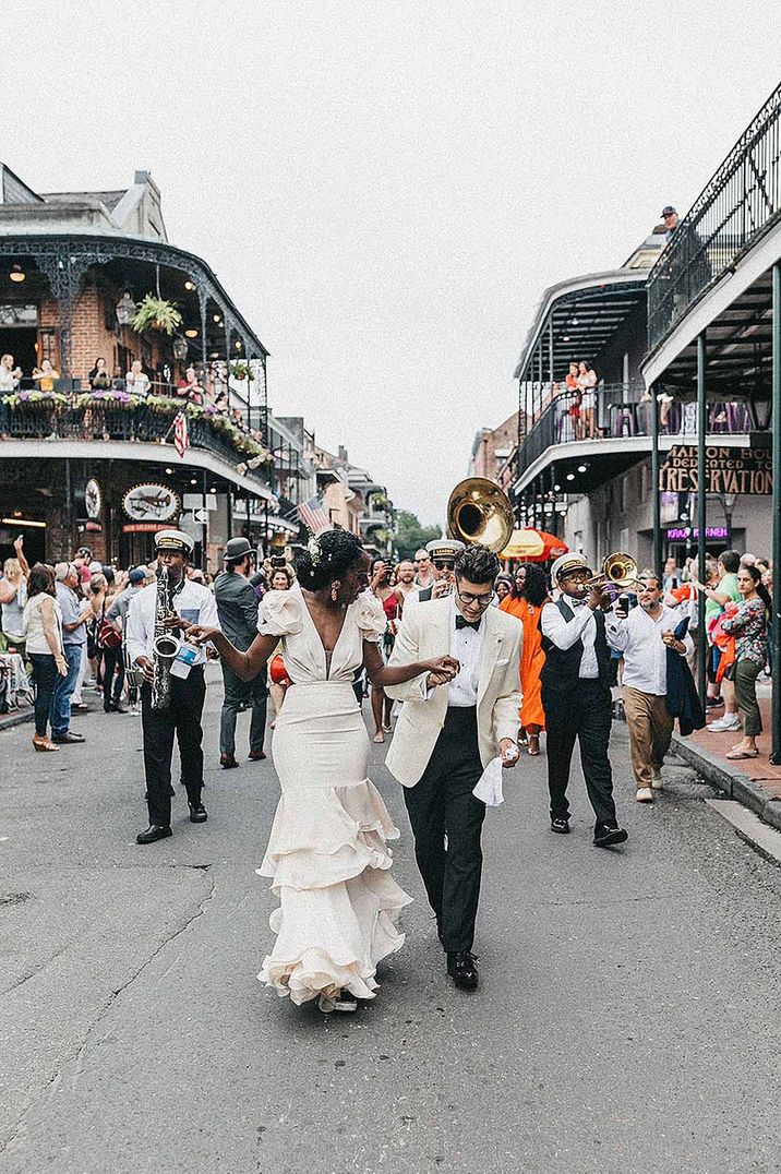 Black bride in a fitted wedding dress with ruffle marching down the street with her groom in a white tuxedo jacket followed by a brass band