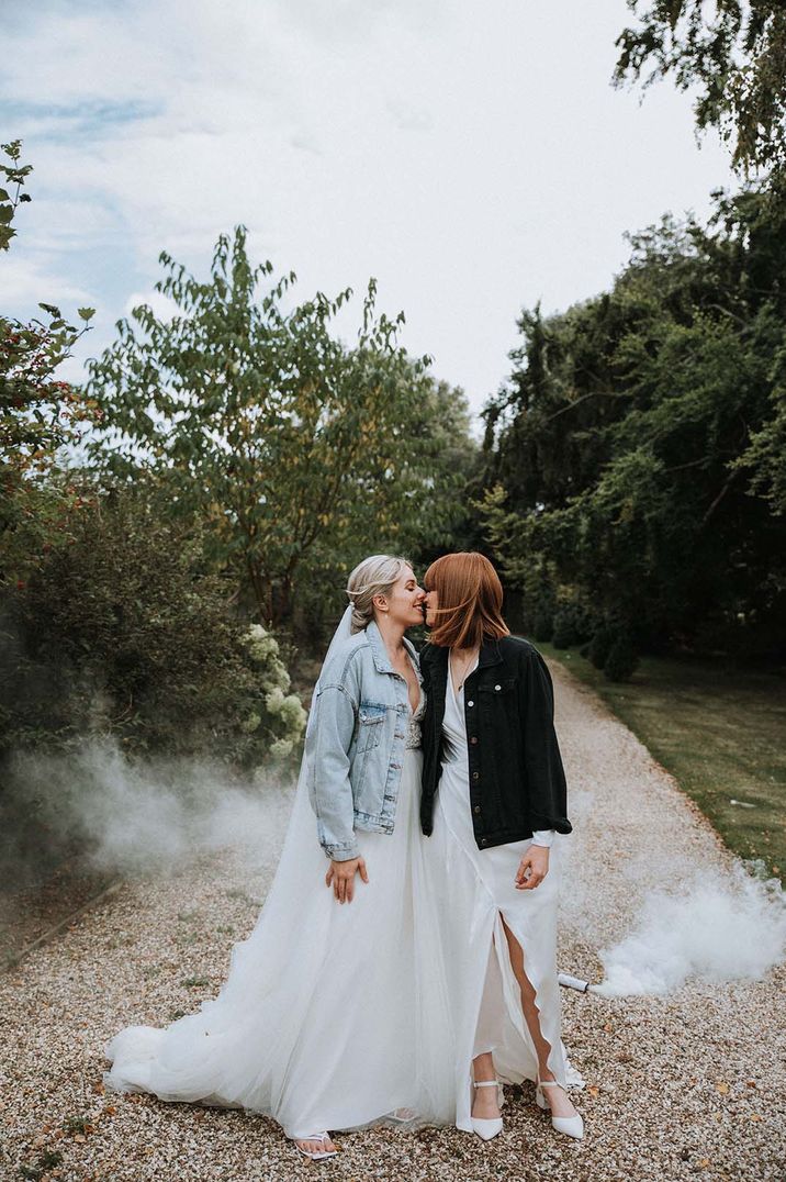 Bride in pale blue denim jacket and bride in black denim jacket kiss with a smoke bomb in the background