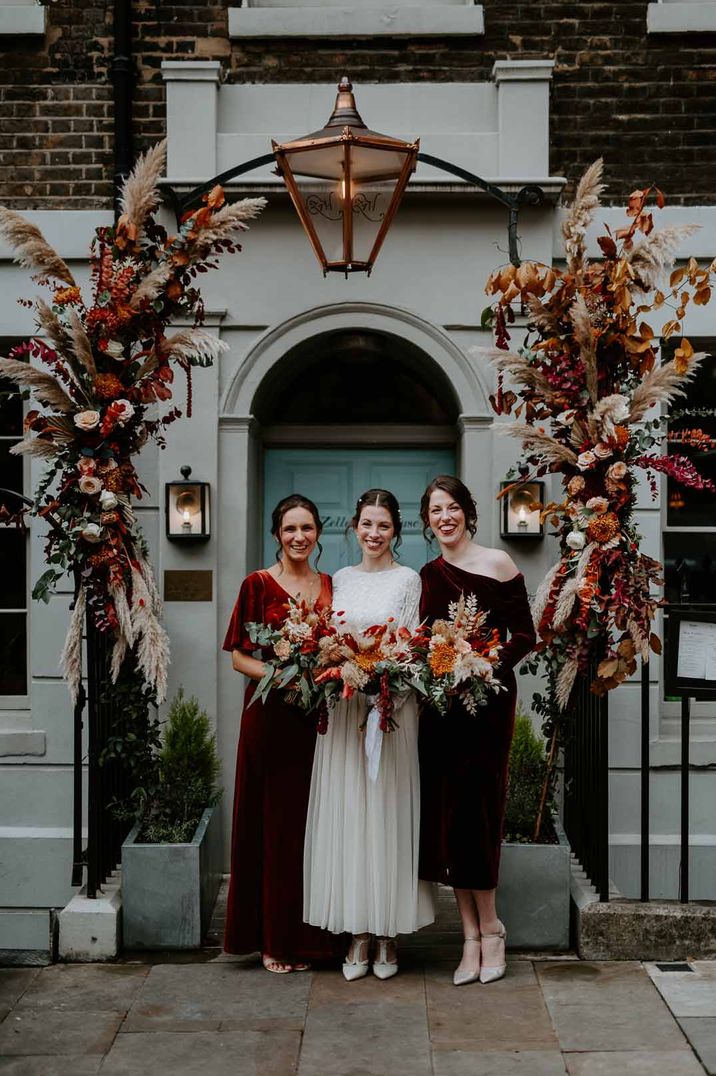 Bride in vintage long sleeve wedding dress standing with bridesmaids in velvet mismatched bridesmaid dresses standing underneath large floral wedding arch outside The Zetter Townhouse London wedding venue 