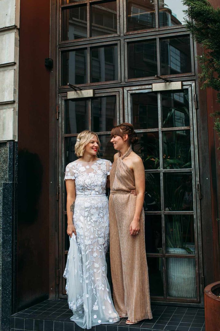 Bride in lace short sleeve wedding dress standing with bridesmaid in gold sparkly halterneck jumpsuit - hen party ideas