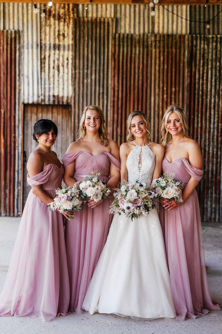 Bride wearing cut out sleeveless lace wedding dress standing with bridesmaids in off the shoulder dusky pink bridesmaid dresses 