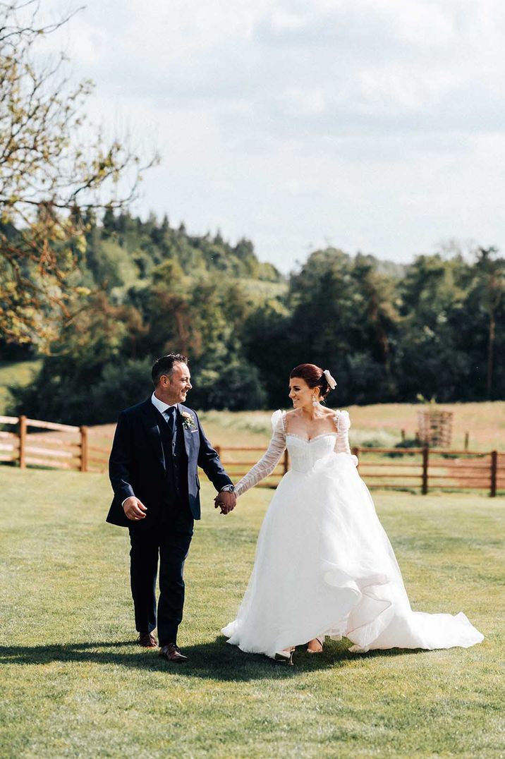 groom holding hands with his bride in a princess tulle wedding dress as they walking through the fields 