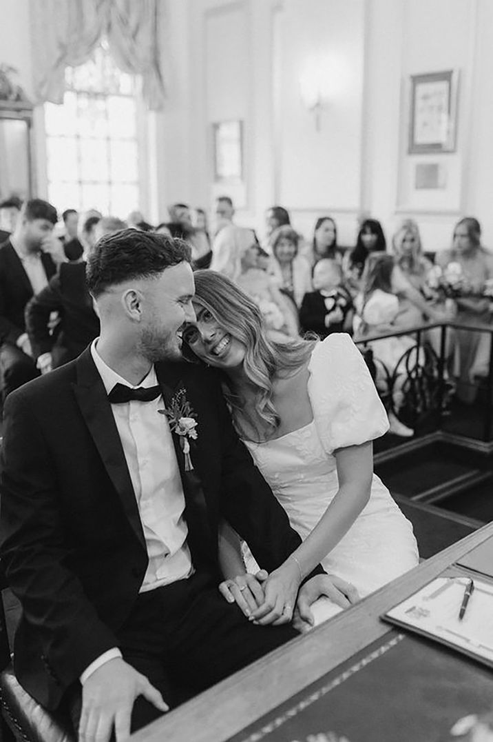 Bride in Story of my Dress wedding dress and groom in black tie embrace as they sign the register at their registry office wedding