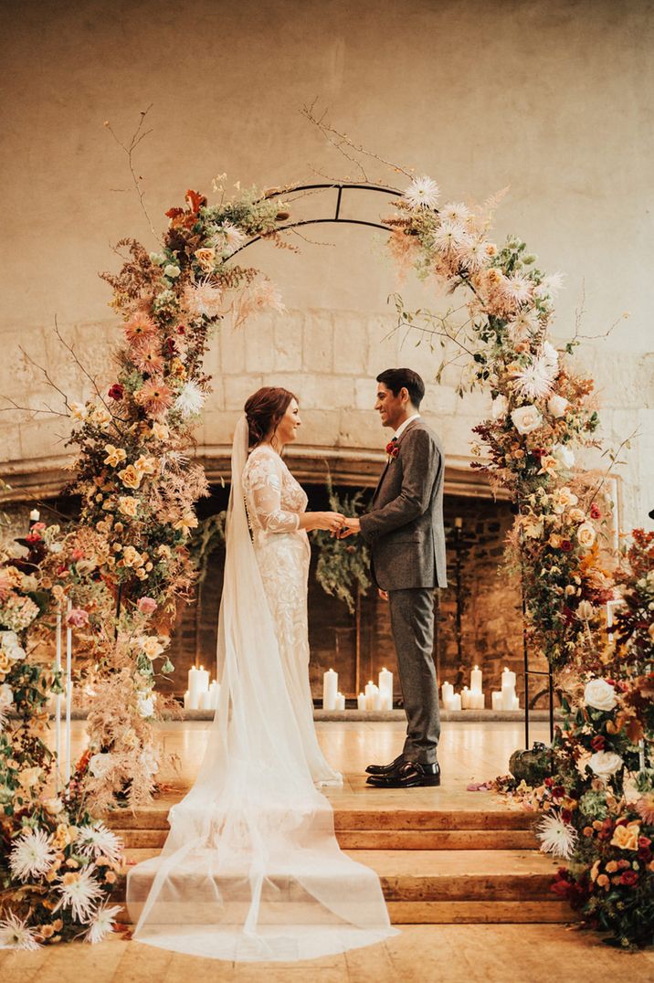 Bride in a lace Hermione de Paula wedding dress exchanging vows with her husband in front of an Autumn floral arch 