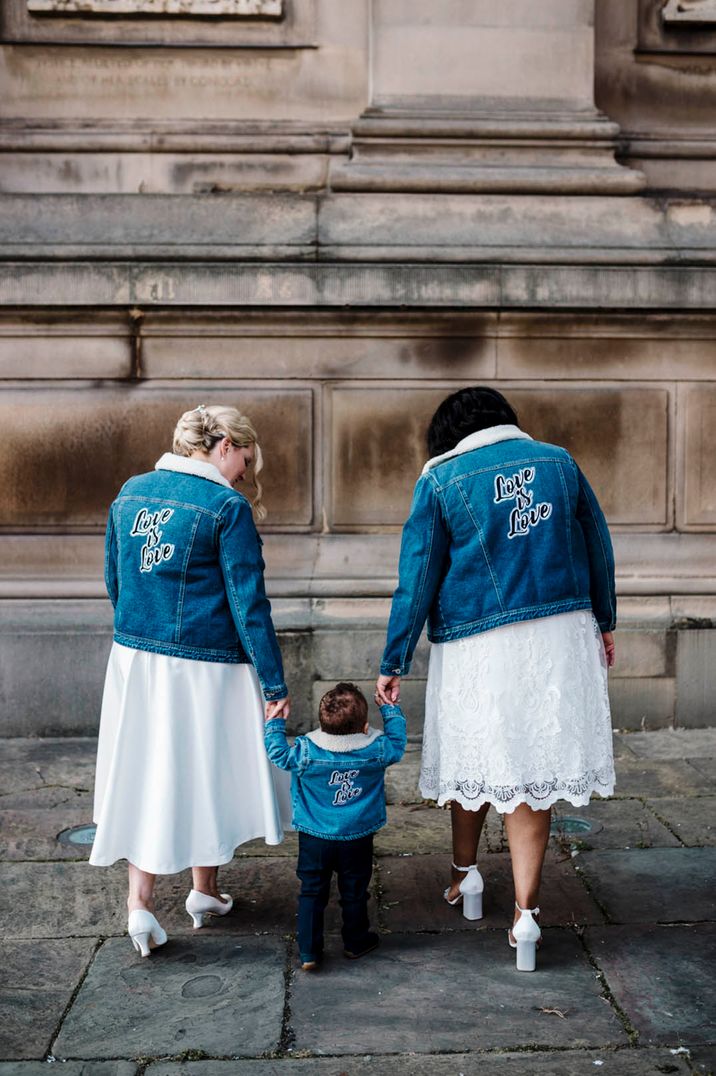 Bride in short wedding dresses wear matching denim jackets with a 'Love is Love' motif with their son 