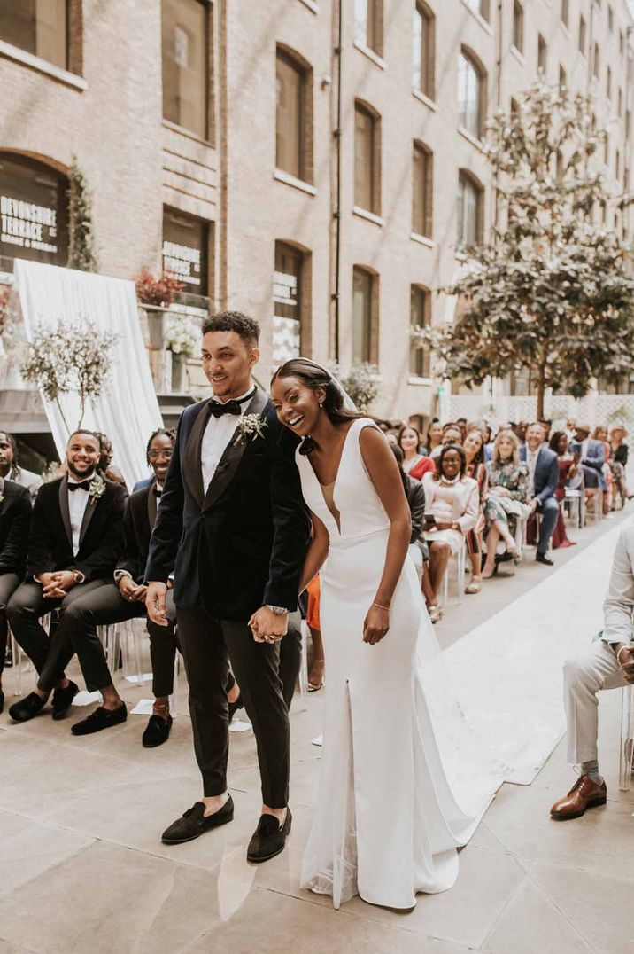 Bride in v-neck sleeveless wedding dress and long train holding hands with groom in classic tux at Devonshire Terrace wedding venue, one of the wedding venues London