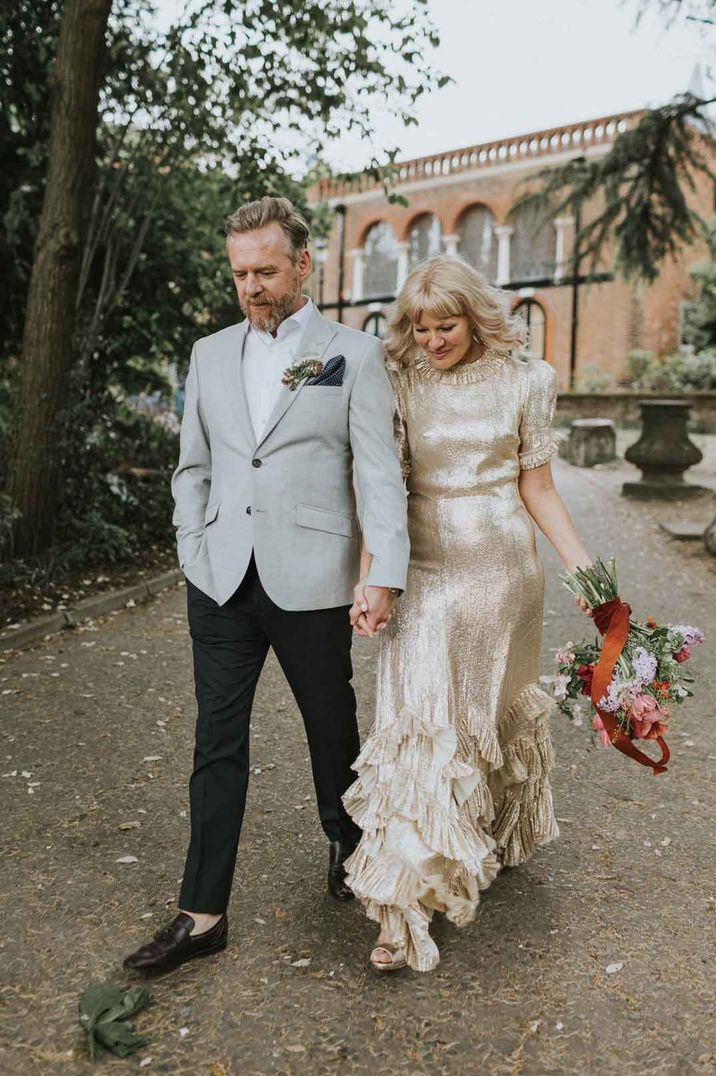 Bride in high neck short sleeve sparkly gold wedding dress with ruffle and layers holding colourful wildflower bridal bouquet walking with groom in grey blazer - how much are wedding dresses