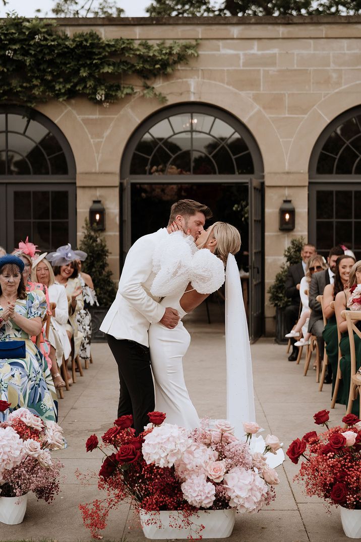 Bride in puff sleeve wedding dress kissing groom in white tuxedo at red and pink theme wedding outdoor ceremony 