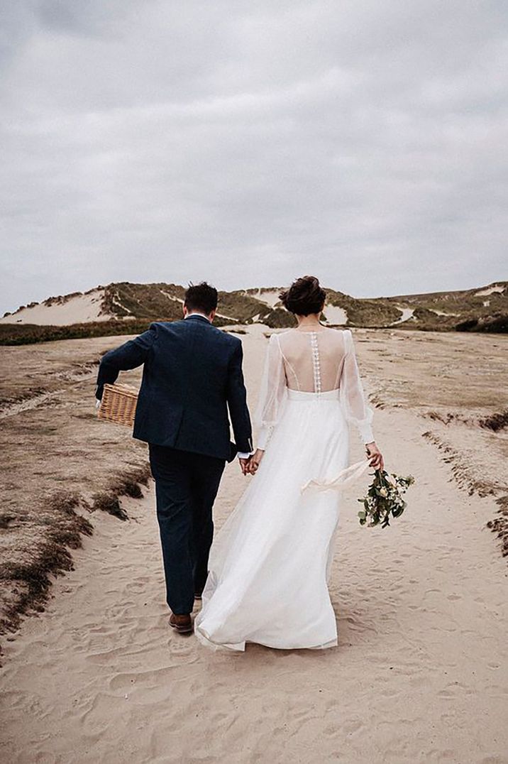 Bride and groom walking hand in hand on Cornwall beach for wedding ceremony with bride wearing a mesh sleeve and open back beach wedding dress