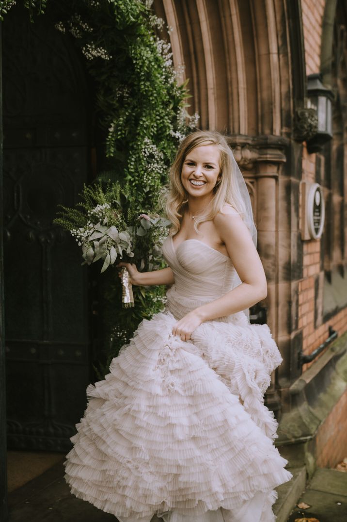 Bride at door of church smiling and holding up the skirt of her blush preloved wedding dress in one hand and bouquet in other