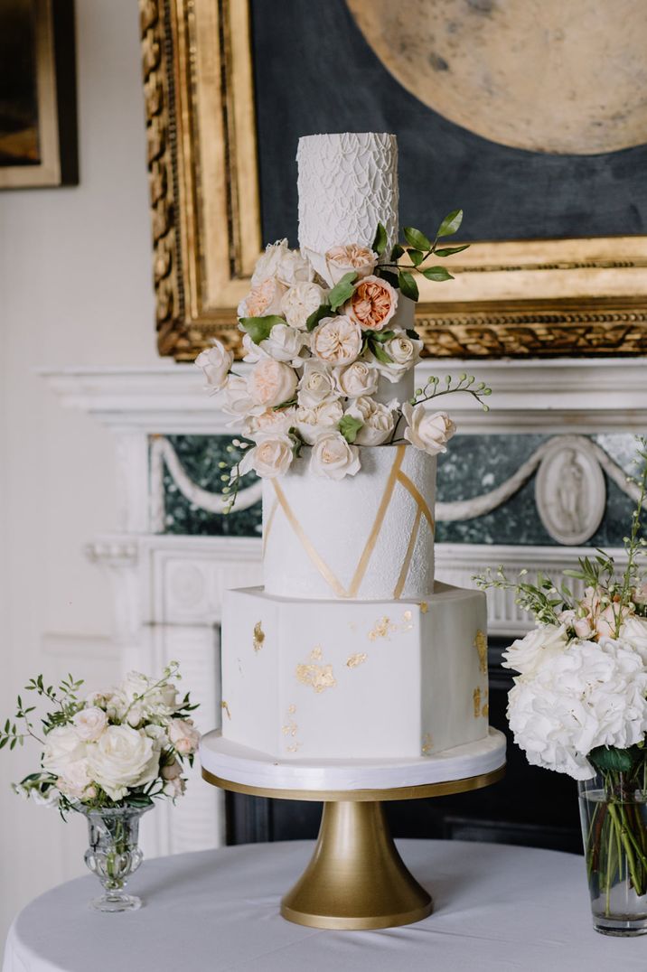 Tall four tier iced wedding cake with hexagonal and round layers decorated with gold leaf and fresh flowers 