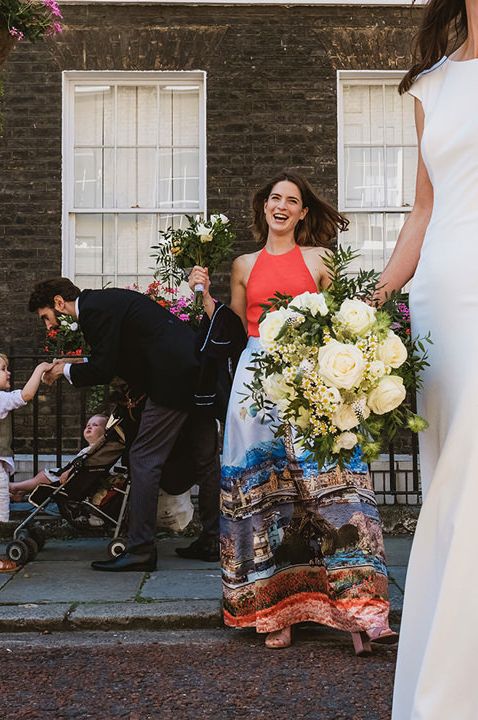 Bride in a fitted wedding dress and lace edge veil holding a white rose bouquet with her bridesmaids in different dresses in the background 