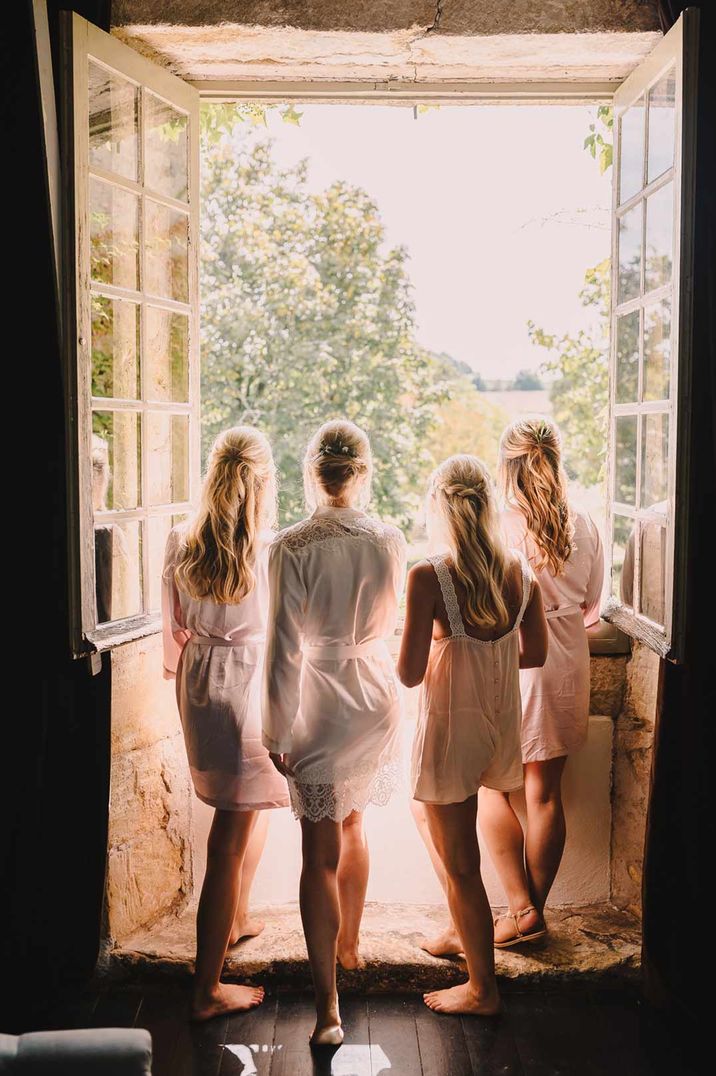 Bride and bridesmaids in satin getting ready robes looking out of window at destination wedding