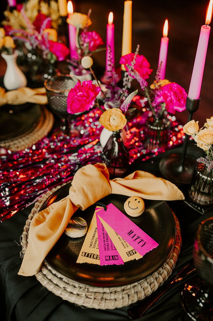 Bright pink and yellow wedding place setting with streamers, smiley face macarons, and bud vases of flowers 