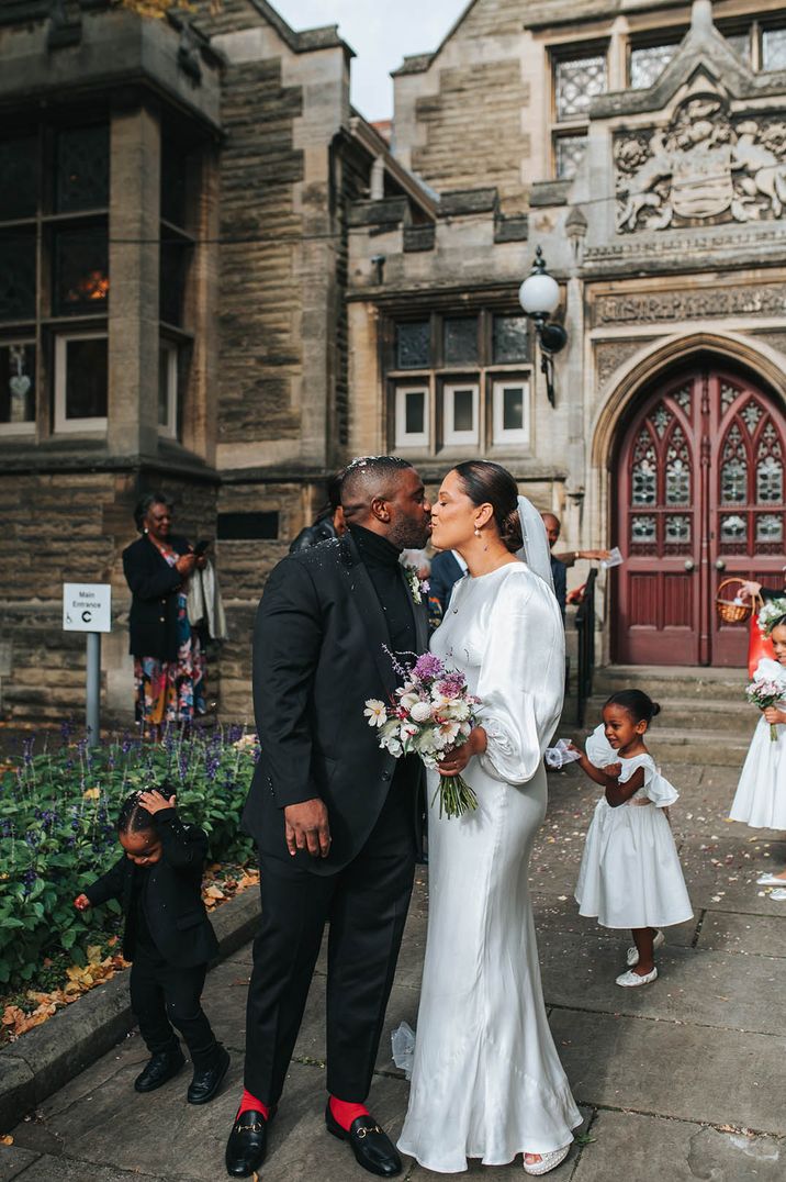 Pregnant bride wearing a long sleeve satin maternity wedding dress kissing the groom for confetti moment 