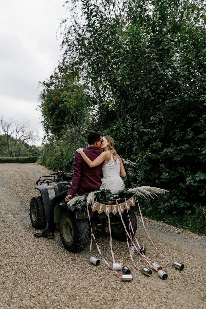 Bride and groom kiss as they ride on a quad bike with tin can decoration 