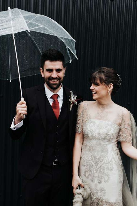 Bride in homemade sage tinted quarter length sleeve wedding dress with homemade bouquet and peacock feathers and groom in dark suit with red tie holding clear umbrellas