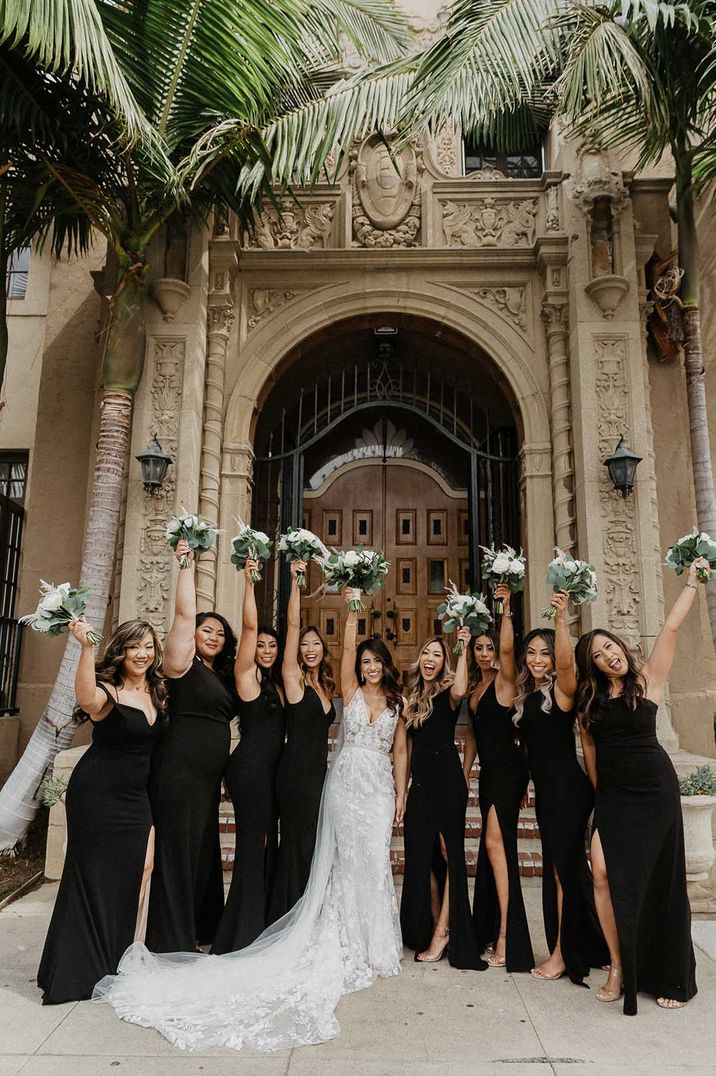 Bridesmaids lift their white floral bouquets in the air and wear long black bridesmaid dresses in differing styles 