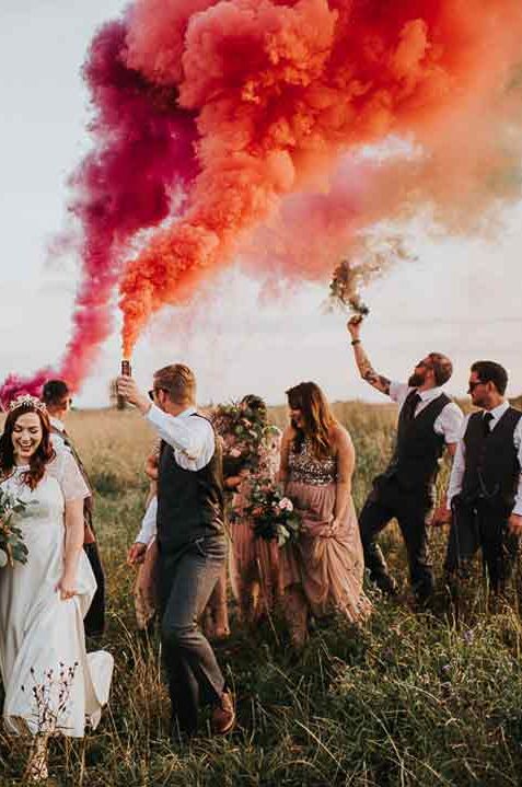 Bride in sheer lace sleeve wedding dress and groom in grey waistcoat and sunglasses walking with wedding party doing colourful orange and pink smoke bomb wedding photos 