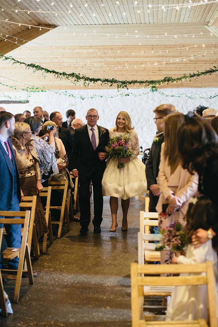 Bride in short long sleeve wedding dress walking down the aisle at Dalduff Luxury Barn Weddings with father of the bride 