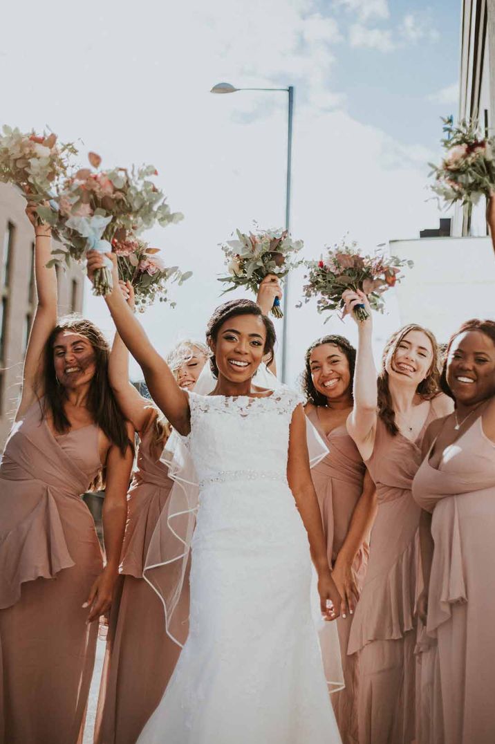 Bride wearing short sleeve boat neck lace wedding dress standing with bridesmaids in neutral ruffle bridesmaid dresses 