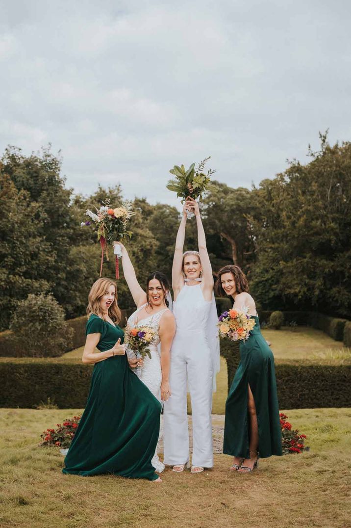 Bride wearing bridal jumpsuit with bridal cape standing with bridesmaids in bottle green velvet bridesmaid dresses - hen party ideas 