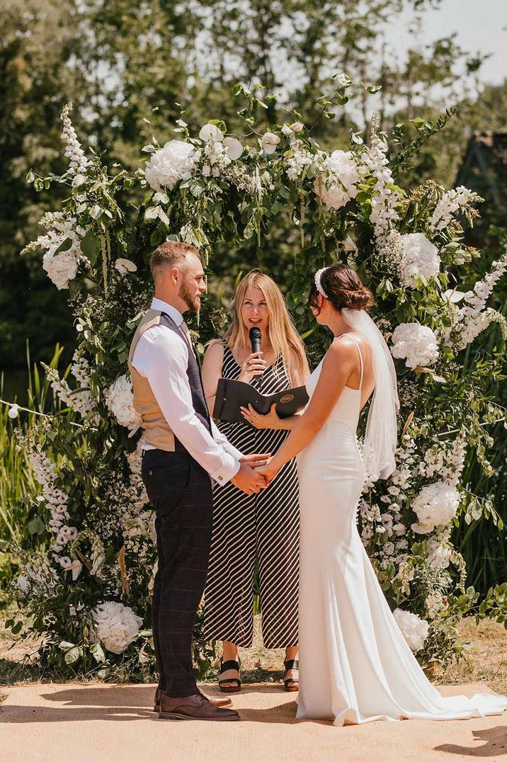 Wedding celebrant in striped jumpsuit stands with bride and groom conducting the outdoor wedding ceremony 