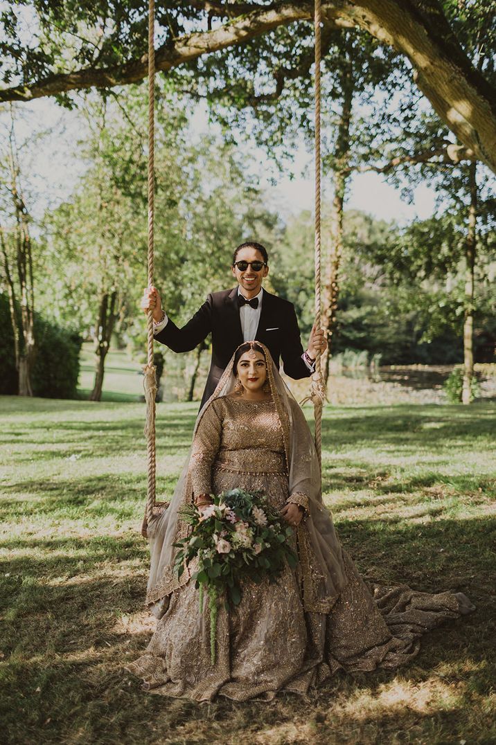 Bride in a gold sari sitting on a swing with her groom in a tuxedo standing behind her 