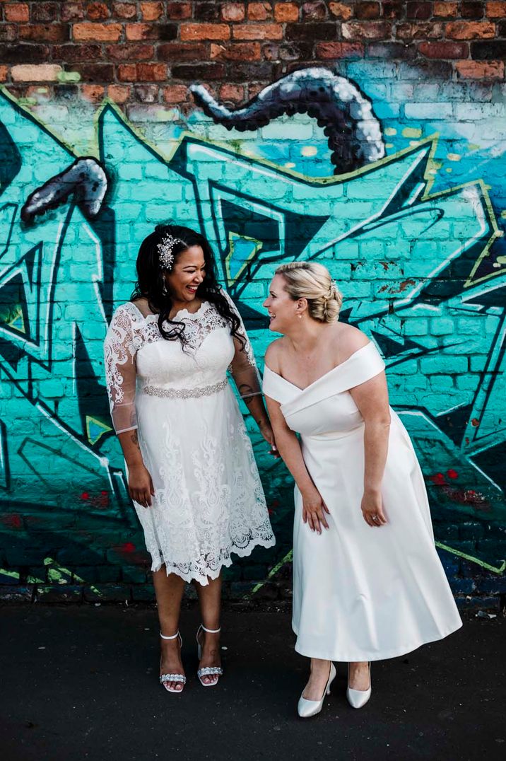 Lesbian wedding with two brides in tea length wedding dresses laughing in front of graffiti art in Liverpool 