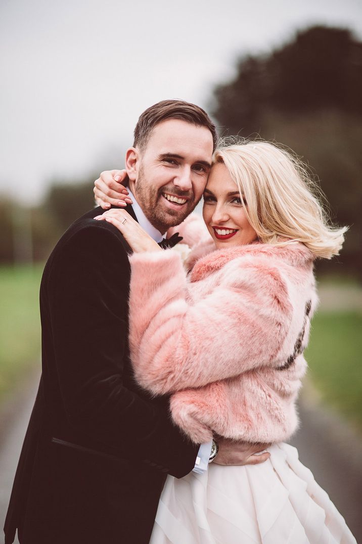 Groom in black tie embraces the bride in a pink faux fur personalised cover up over her white strapless dress