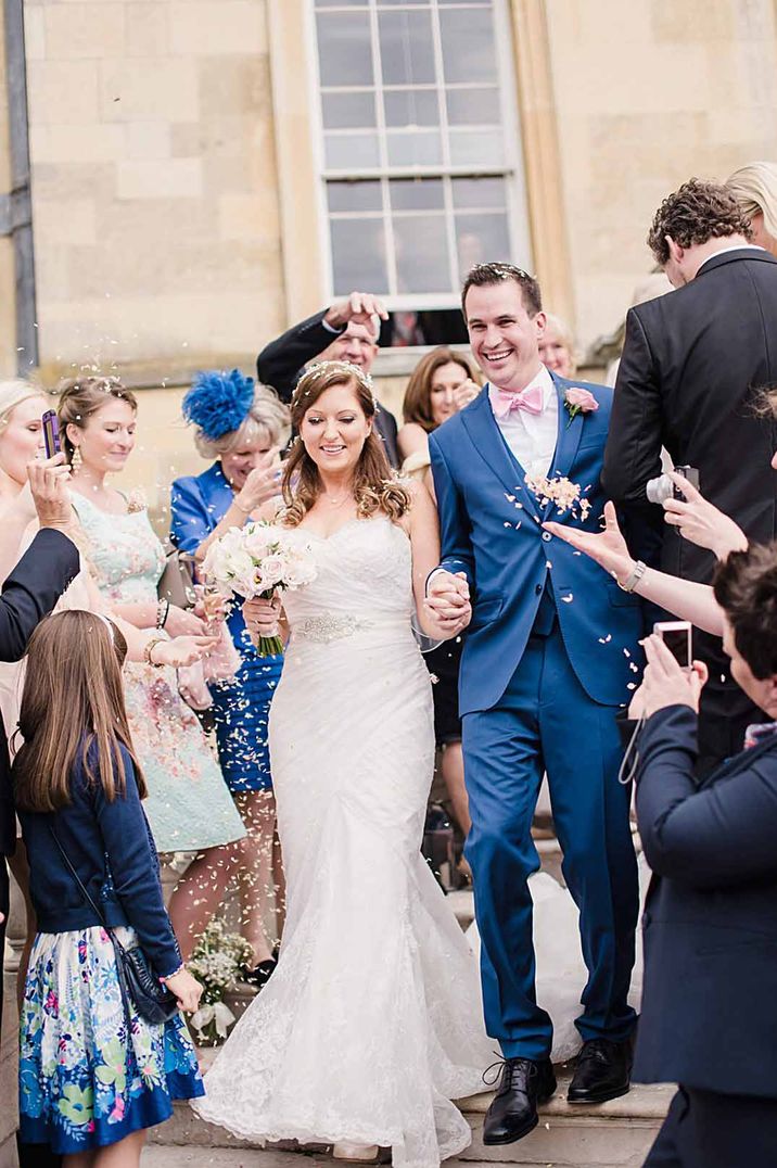 Bride in strapless peplum wedding dress doing confetti walk with groom in blue suit and pink bowtie 