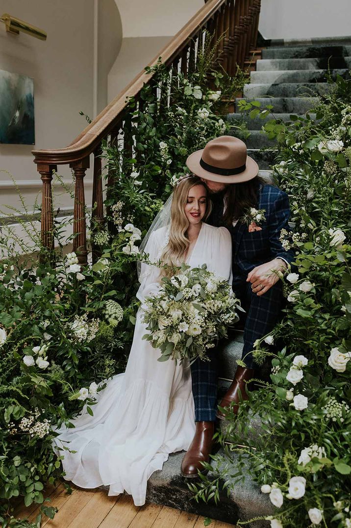 Bride in v-neck long sleeve puff wedding dress with groom in alternative checkered suit and hat sitting on the stairs of Netherbyres House Scottish wedding venue surrounded by white flowers and foliage arrangements 