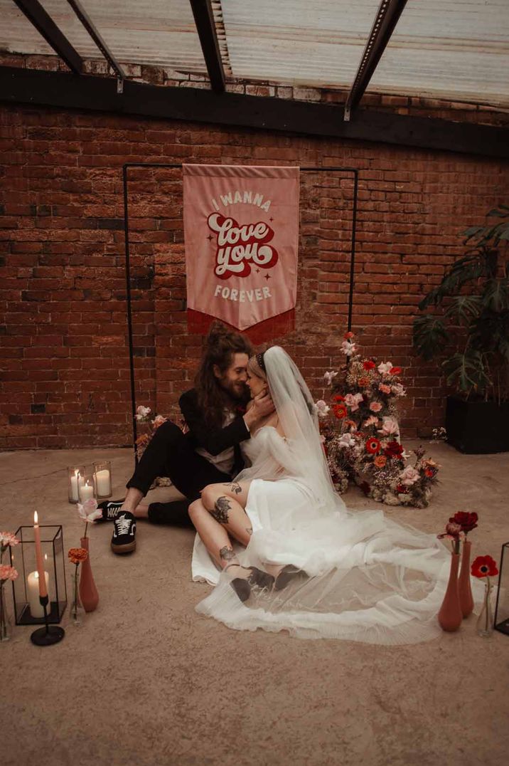 Bride in short wedding dress and cathedral length veil sitting with groom in alternative grooms suit for Valentines-inspired wedding with large pink fabric wedding banner at The Shack Revolution wedding venue