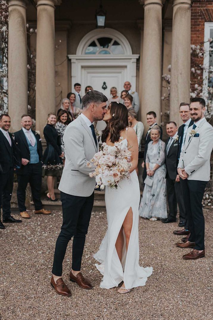 Bride in satin slip wedding dress with split leg kissing groom in grey suit 