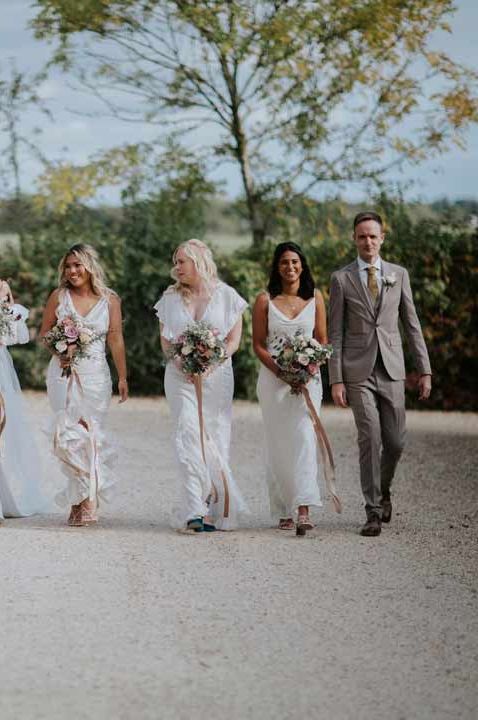 Bride walks with her mixed gender wedding party and bridesmate in white dresses and a grey suit