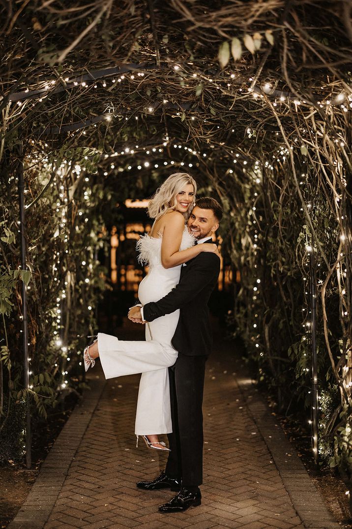 The groom in traditional black tie lifts the bride wearing a feather Nadine Merabi wedding jumpsuit 