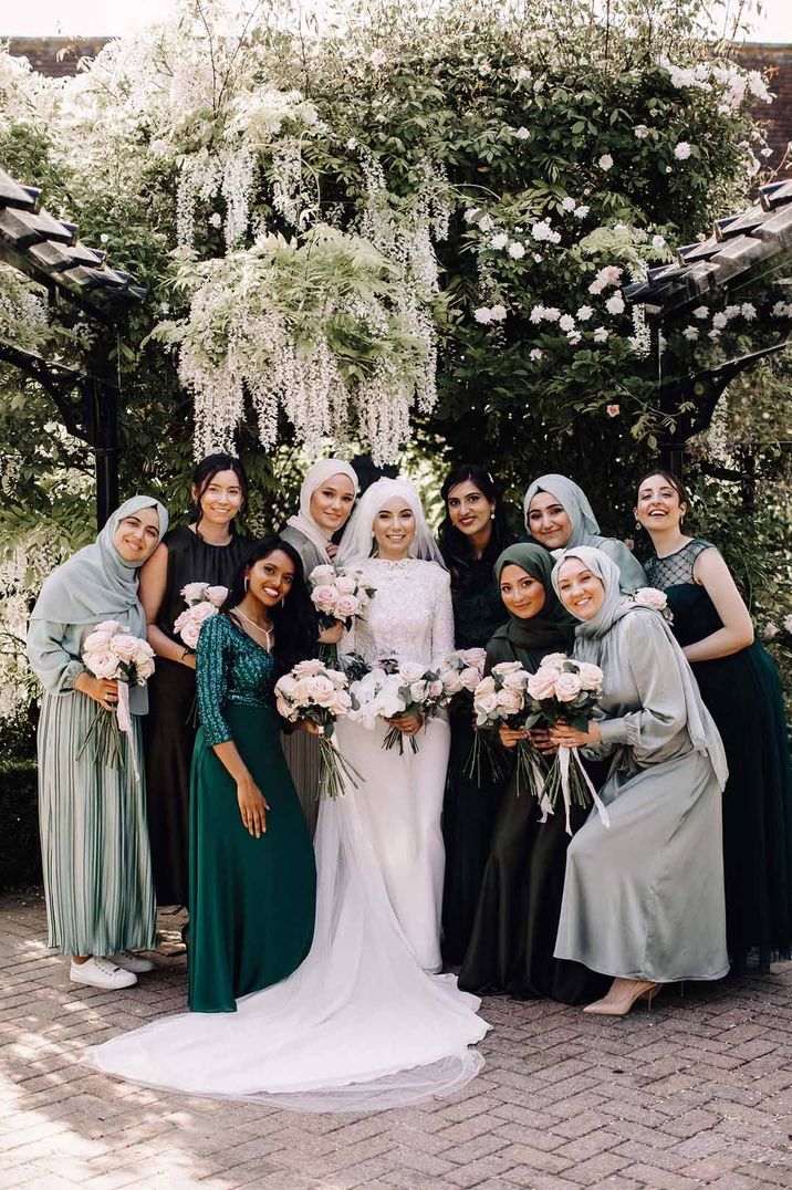 Bride in long sleeve wedding dress standing with bridal party in mismatched green bridesmaid dresses 