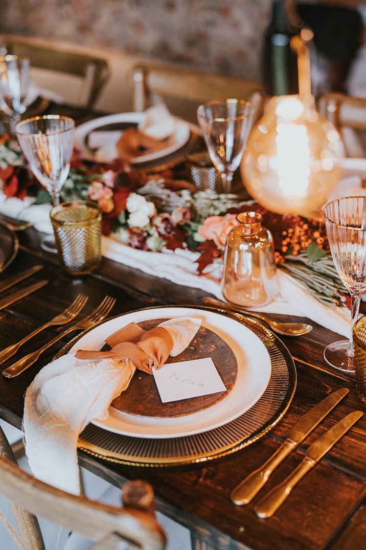 Metallic charger plates with white and orange napkin, rustic place name cards, gold rimmed glasses and autumnal floral table arrangements 