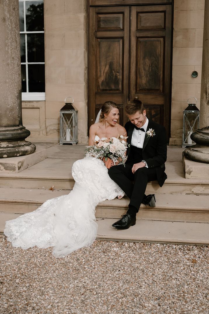 Groom in black tuxedo laughing with the bride in a lace wedding dress on the steps at the country house wedding venue 