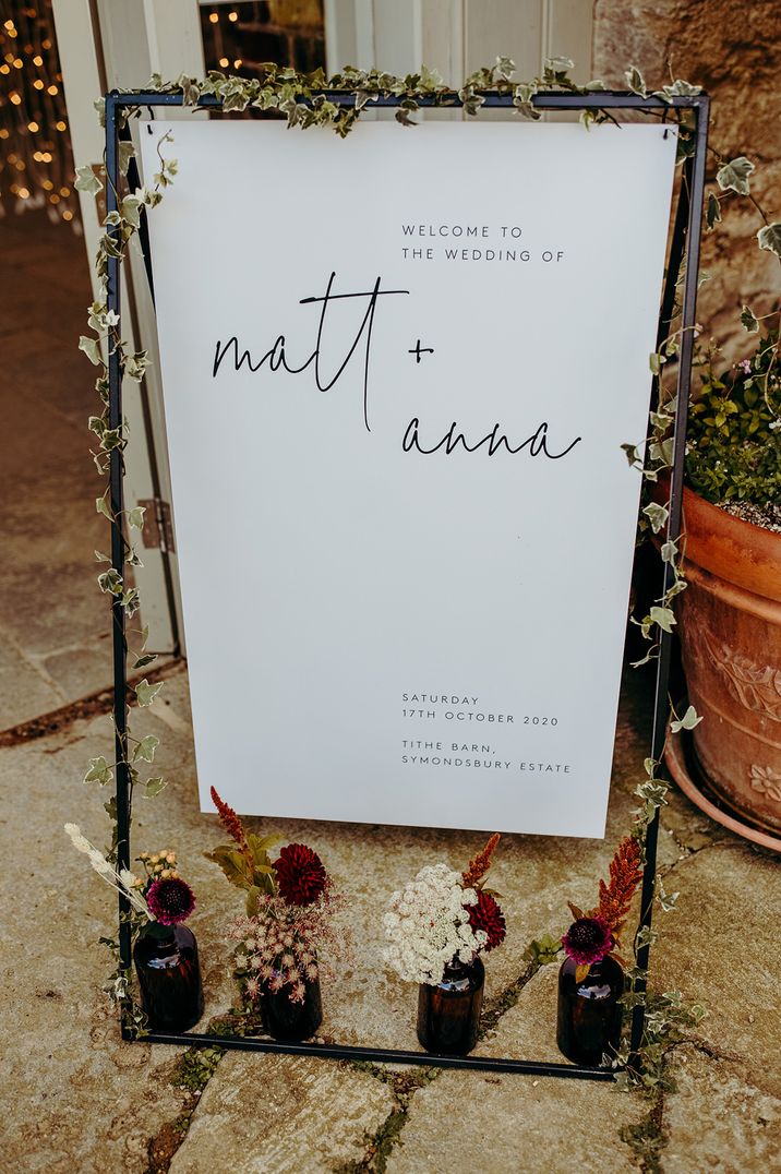 White wedding welcome sign on a black stand decorated with ivy and autumnal flowers 